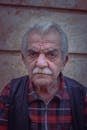 Close-up portrait of an elderly man in a plaid shirt and leather vest against a textured wall.